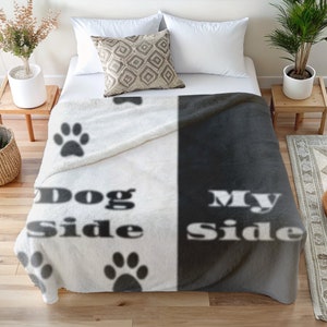 Könnte beinhalten: Zweifarbige Decke, weiß und dunkelgrau. Die weiße Seite hat schwarze Pfotenabdrücke und die Worte "Dog Side", die graue Seite sagt "My Side". Die Decke liegt auf einem Bett mit Kissen und Pflanzen. Ein gemütliches Wohnaccessoire.