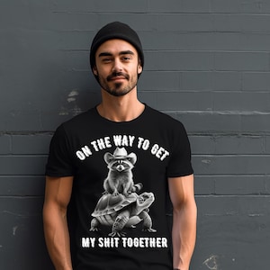 Könnte beinhalten: Schwarzes T-Shirt mit dem Text "ON THE WAY TO GET MY SHIT TOGETHER". Die Grafik zeigt einen Waschbären mit Cowboyhut, der auf einer Schildkröte reitet. Die Person auf dem Bild trägt eine schwarze Mütze.