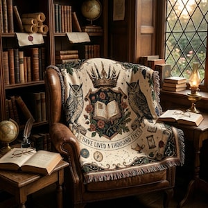 Könnte beinhalten: Ein gemütliche Leseecke mit einem braunen Ledersessel und einer cremefarbenen Decke. Die Decke zeigt Eulen, eine Krone und den Text "I HAVE LIVED A THOUSAND LIVES". Bücherregale und ein Fenster vervollständigen die Szene.