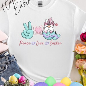 Könnte beinhalten: Weißes T-Shirt mit einem farbenfrohen Oster-Motiv. Das Design zeigt ein Peace-Zeichen, ein Herz und einen Zwerg, der aus einem Ei schlüpft, mit den Worten "Peace Love Easter". Die Worte "Happy Easter" sind kursiv geschrieben.