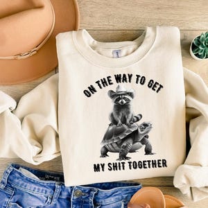 Könnte beinhalten: Cremefarbenes Sweatshirt mit dem Text "ON THE WAY TO GET MY SHIT TOGETHER." Die Grafik zeigt einen Waschbären mit Cowboyhut, der auf einer Schildkröte reitet. Das Design ist in Schwarz und Weiß.