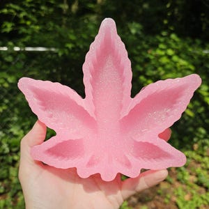 May include: A pink, glittery cannabis leaf-shaped tray. The tray is held in a hand, and the background is blurred green foliage. The tray is a light pink color and has a sparkly, glittery finish.