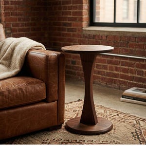 May include: A dark brown, wooden side table with a round top and a unique hourglass-shaped base. The table is set on a patterned rug next to a brown leather sofa. A beige blanket with fringe is draped over the sofa.