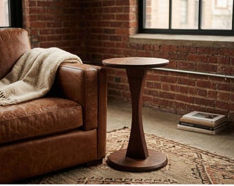 May include: A dark brown, wooden side table with a round top and a unique hourglass-shaped base. The table is set on a patterned rug next to a brown leather sofa. A beige blanket with fringe is draped over the sofa.