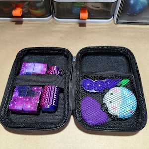May include: Open black zipped case containing various items. Includes two galaxy-themed cubes, a purple cylinder, a purple textured item, a blue and white ball, and a purple beaded item. The case is open, revealing the contents.