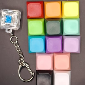 May include: A selection of multi-coloured square keycaps in shades of red, orange, yellow, green, blue, purple, pink, and black. A clear, square keychain with a blue design is attached to a silver chain.
