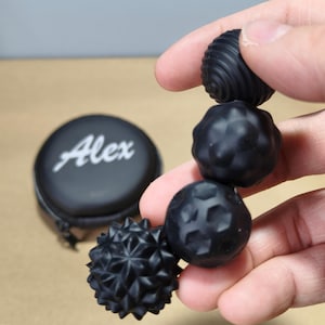 May include: A hand holds a set of black massage balls with different textured surfaces. A black case with the name "Alex" in white lettering is visible. The balls are likely made of rubber or a similar material.
