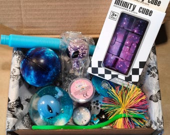 May include: A sensory toy gift box containing a blue pop tube, a galaxy-patterned infinity cube in packaging, a blue galaxy ball, a clear slime container, a small alien figure, a blue alien-filled water ball, and a multi-colored spiky rubber ball.