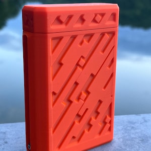 May include: Bright orange rectangular case with a geometric design. The case has a textured surface with diagonal lines and angular shapes. The top of the case has a series of small, repeating geometric patterns. The case is sitting on a concrete surface.