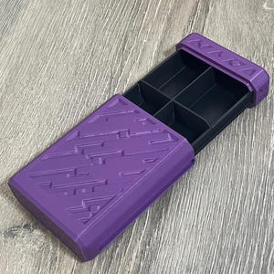 May include: A purple rectangular box with a sliding drawer, featuring a geometric pattern on the lid. The interior of the box is divided into six compartments. The box is open, revealing the black interior.