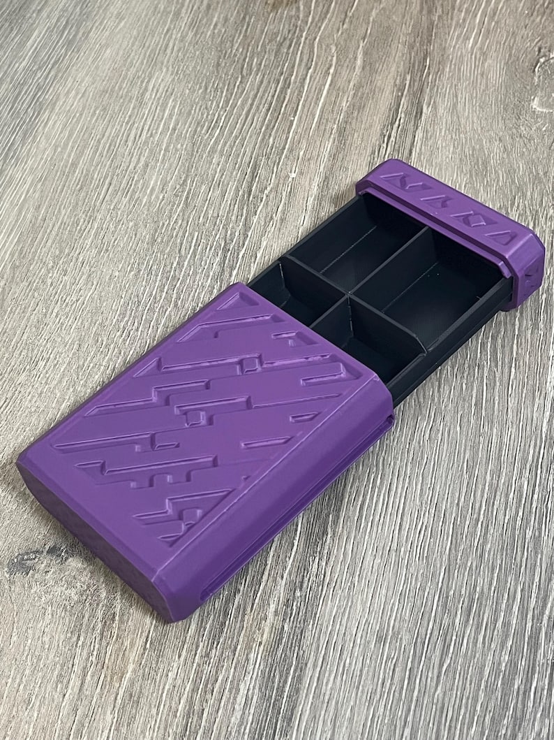 May include: A purple rectangular box with a sliding drawer, featuring a geometric pattern on the lid. The interior of the box is divided into six compartments. The box is open, revealing the black interior.