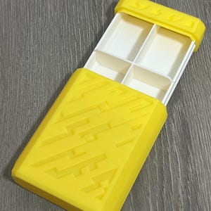 May include: A yellow rectangular pill organizer with a sliding lid. The lid and base are yellow with a geometric design. The interior has four white compartments for storing pills or small items. The organizer is open, revealing the compartments.
