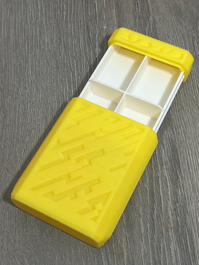 May include: A yellow rectangular pill organizer with a sliding lid. The lid and base are yellow with a geometric design. The interior has four white compartments for storing pills or small items. The organizer is open, revealing the compartments.
