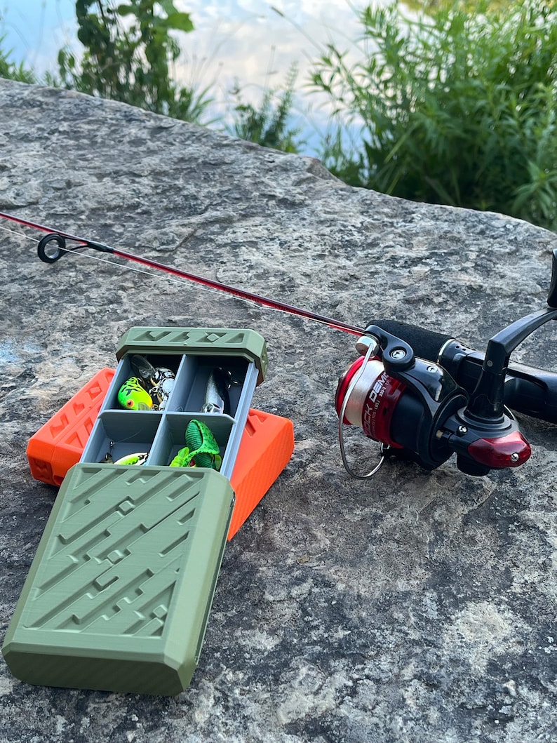 May include: An open green fishing tackle box with various lures sits on a rock next to an orange base. A fishing rod and reel are positioned to the right. The lures are in different colors, including green, silver, and yellow. The background shows water and greenery.