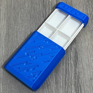 May include: A blue plastic organizer with a sliding lid. The lid and base are a vibrant blue, while the interior features four white compartments. The lid has a textured design and a series of small, triangular shapes.