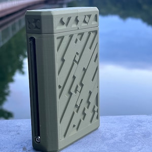 May include: A rectangular, olive-green case with a geometric pattern on the front and top. The case has a small, clear window on the side and a metal accent. The background is a blurred view of a lake.