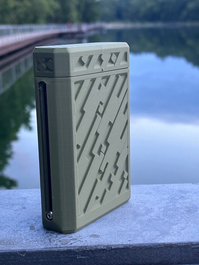May include: A rectangular, olive-green case with a geometric pattern on the front and top. The case has a small, clear window on the side and a metal accent. The background is a blurred view of a lake.