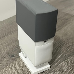 May include: A modern, two-tone soap dispenser. The top is a solid, matte gray, while the body is white. The dispenser has a rectangular base and a curved, rounded body. The design is minimalist and sleek.