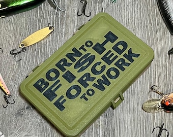 May include: An olive green fishing tackle box with the text "BORN TO FISH, FORCED TO WORK" in black. Various fishing lures and hooks surround the box, including green, gold, and silver lures. The background is a light gray wood grain.
