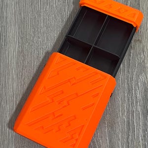 May include: An open, orange rectangular box with a black interior divided into four compartments. The box has a textured design on the front and the word "KABBA" is visible on the top. The box is on a gray wooden surface.