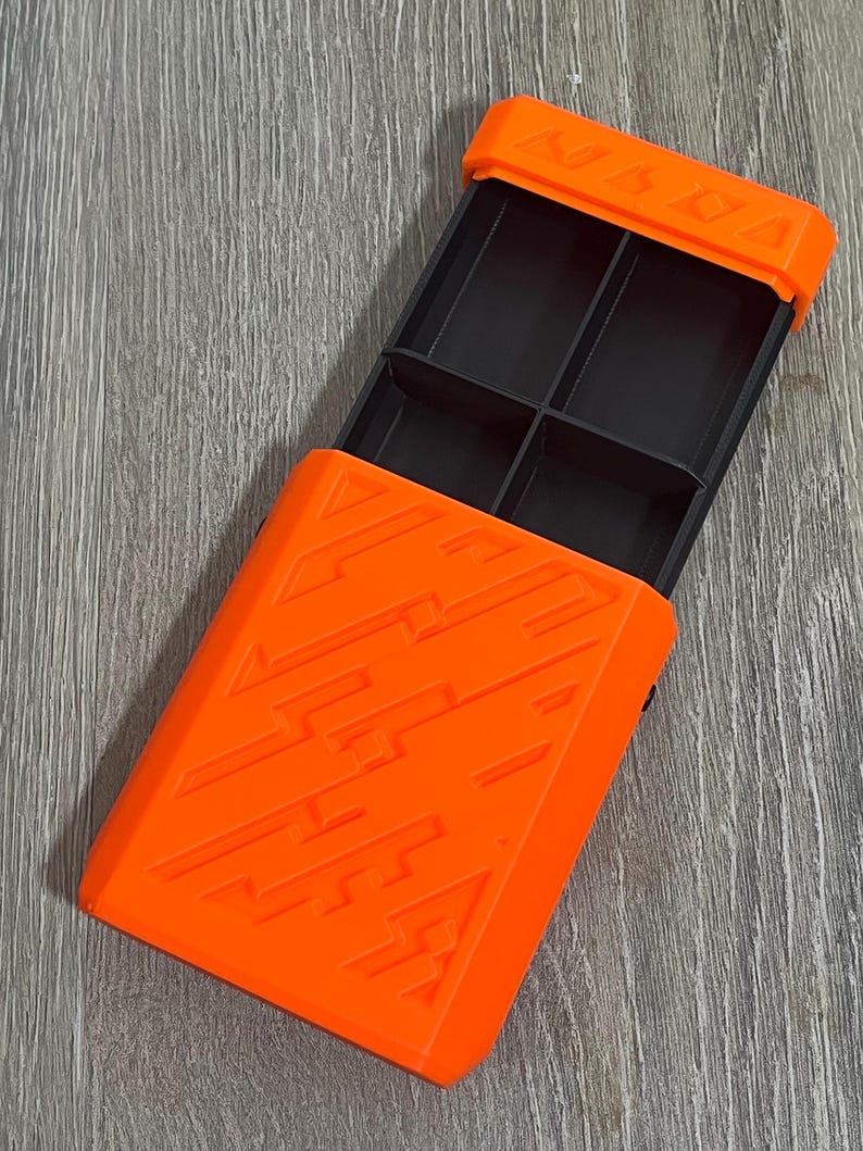 May include: An open, orange rectangular box with a black interior divided into four compartments. The box has a textured design on the front and the word "KABBA" is visible on the top. The box is on a gray wooden surface.
