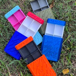 May include: A collection of colorful, rectangular storage containers with divided compartments. The containers are in various colors, including pink, blue, orange, and green, with contrasting colored lids. The containers are arranged on a bed of green grass.
