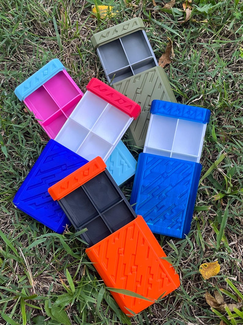 May include: A collection of colorful, rectangular storage containers with divided compartments. The containers are in various colors, including pink, blue, orange, and green, with contrasting colored lids. The containers are arranged on a bed of green grass.