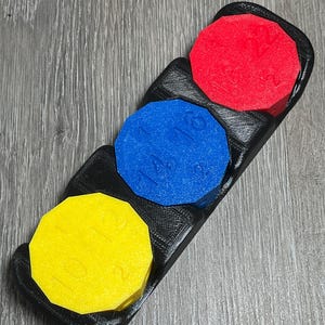May include: A black 3D-printed organizer holding three colorful, octagonal tokens. The tokens are red, blue, and yellow, each with a number printed on them. The organizer sits on a gray wooden surface.