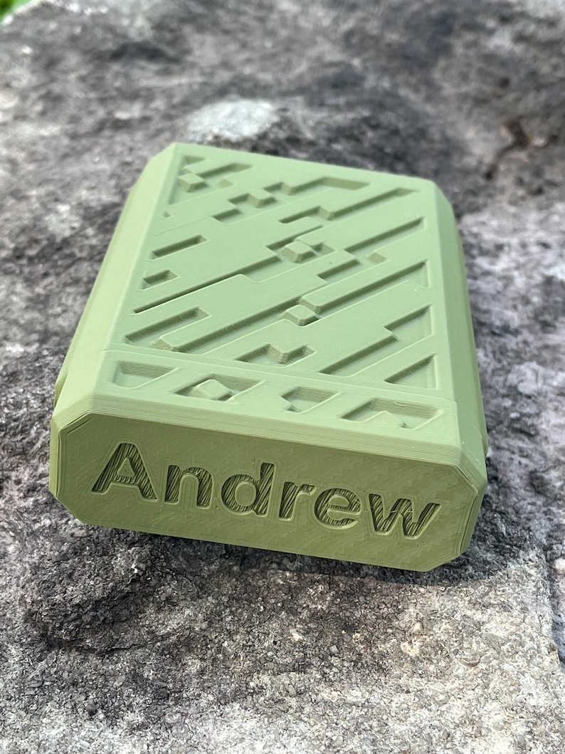May include: A rectangular, olive-green case with a geometric pattern on the top surface. The name "Andrew" is engraved on the front. The case is resting on a textured, gray stone surface.
