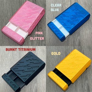 May include: Four small rectangular boxes in pink glitter, clear blue, burnt titanium, and gold. Each box has a sliding drawer with a white interior. The boxes have a geometric design on the top and sides, and the text "V/S/S/A" is visible on the drawer.
