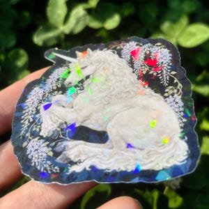 Puede incluir: Una pegatina hologr&aacute;fica con un unicornio blanco con un cuerno en espiral, descansando entre flores de glicina azules y blancas. La pegatina tiene un borde azul oscuro y acentos iridiscentes, creando un dise&ntilde;o de fantas&iacute;a.