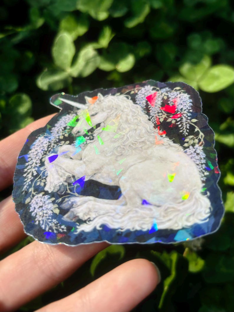 Puede incluir: Una pegatina hologr&aacute;fica con un unicornio blanco con un cuerno en espiral, descansando entre flores de glicina azules y blancas. La pegatina tiene un borde azul oscuro y acentos iridiscentes, creando un dise&ntilde;o de fantas&iacute;a.
