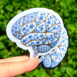 May include: A die-cut sticker shaped like a human brain, featuring a blue and white floral pattern reminiscent of porcelain, with gold-colored lines separating the sections of the brain. The design includes intricate geometric and botanical motifs.