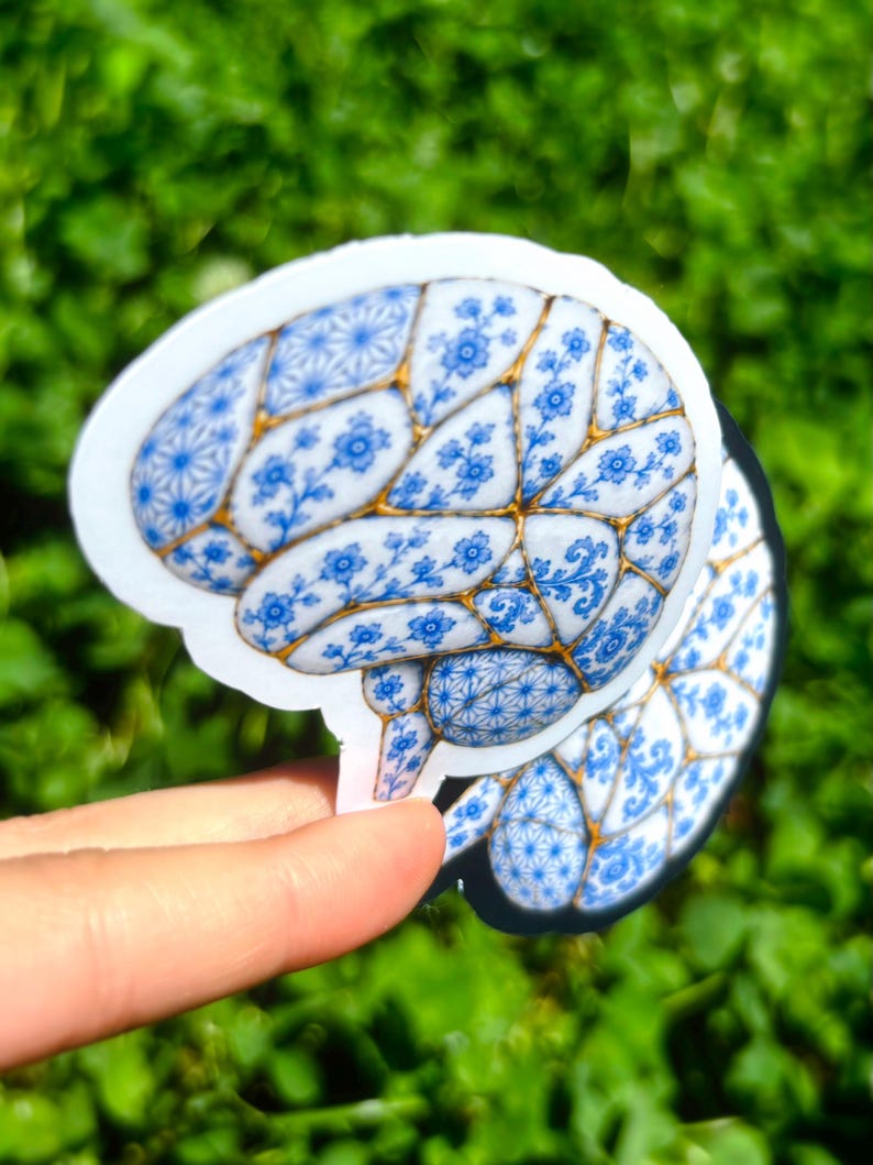 May include: A die-cut sticker shaped like a human brain, featuring a blue and white floral pattern reminiscent of porcelain, with gold-colored lines separating the sections of the brain. The design includes intricate geometric and botanical motifs.