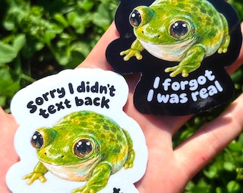 以下が含まれることがあります： 大きな暗い目をした緑色のカエルが描かれた2枚のダイカットステッカー。テキストは「Sorry I didn't text back, I forgot I was real」と書かれています。1枚は黒い背景、もう1枚は白い背景で、どちらも同じカエルのイラストが描かれています。