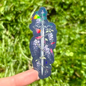 May include: A holographic sticker featuring a sword design with wisteria flowers and red accents. The sticker has a dark background and a rainbow effect, with a silver sword in the center.