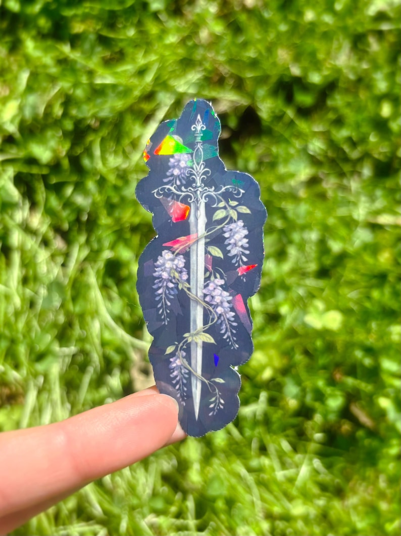 May include: A holographic sticker featuring a sword design with wisteria flowers and red accents. The sticker has a dark background and a rainbow effect, with a silver sword in the center.