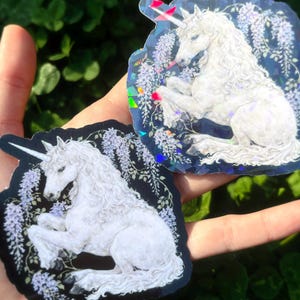 Puede incluir: Dos pegatinas con un unicornio blanco con una melena fluida y un cuerno en espiral. El unicornio est&aacute; sobre un fondo oscuro con flores moradas. Una pegatina tiene un fondo azul y la otra un fondo negro.
