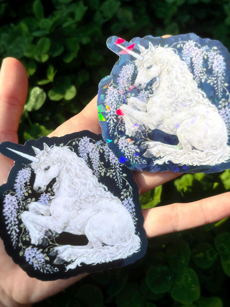 Puede incluir: Dos pegatinas con un unicornio blanco con una melena fluida y un cuerno en espiral. El unicornio est&aacute; sobre un fondo oscuro con flores moradas. Una pegatina tiene un fondo azul y la otra un fondo negro.