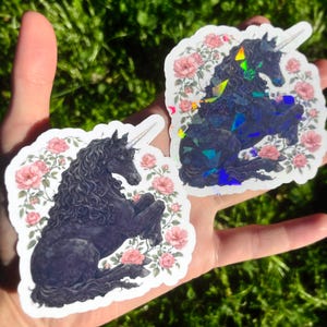 May include: Two unicorn stickers. One is a matte black unicorn with a white horn and surrounded by pink roses. The other is a holographic unicorn with a white horn and surrounded by pink roses. The stickers are held in a hand against a green background.