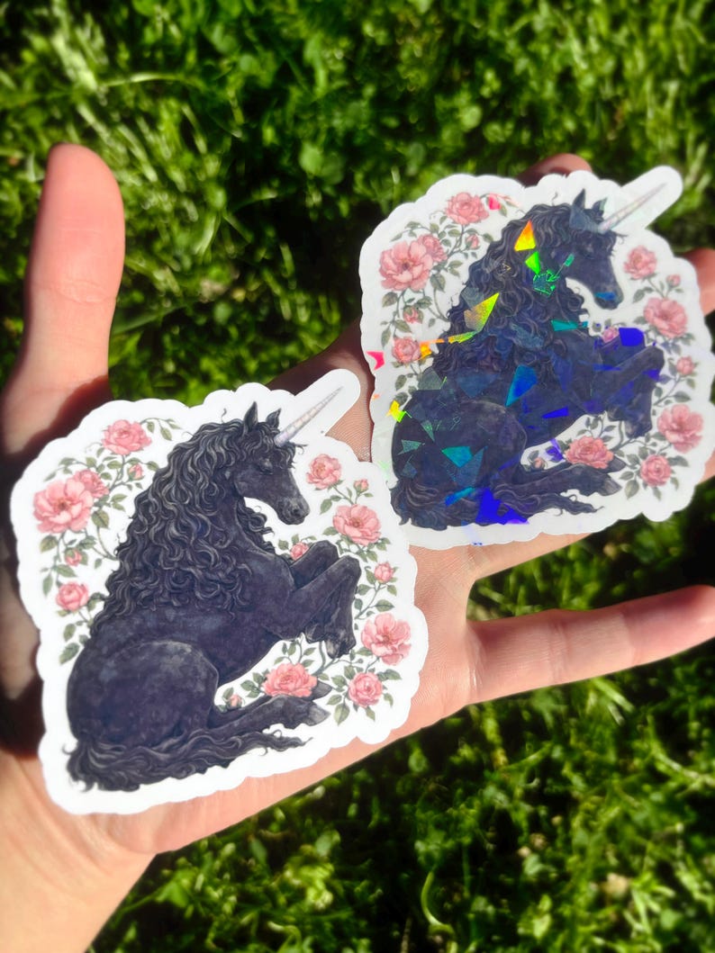 May include: Two unicorn stickers. One is a matte black unicorn with a white horn and surrounded by pink roses. The other is a holographic unicorn with a white horn and surrounded by pink roses. The stickers are held in a hand against a green background.