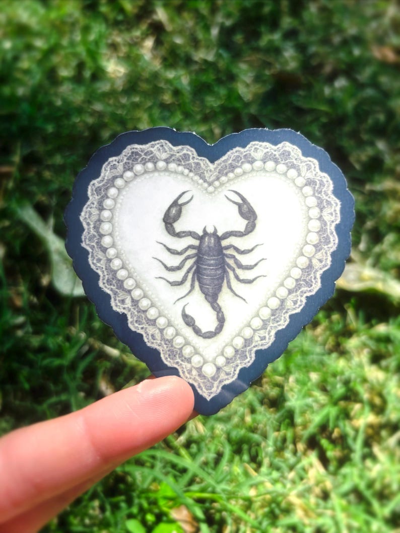 Pu&ograve; includere: Un adesivo a forma di cuore con un'illustrazione dettagliata di uno scorpione. Il design presenta uno sfondo bianco, un bordo di perle e un bordo smerlato blu navy. L'adesivo &egrave; tenuto su uno sfondo verde sfocato.