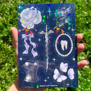 May include: A dark blue sticker sheet with holographic accents featuring various illustrations. Designs include a white flower, a sword with wings, a corset, butterflies, a tooth, a helmet, and a bow. The text "PEACH CAT PRINTS" is visible.