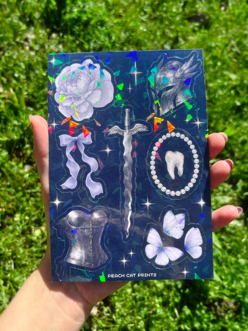 May include: A dark blue sticker sheet with holographic accents featuring various illustrations. Designs include a white flower, a sword with wings, a corset, butterflies, a tooth, a helmet, and a bow. The text "PEACH CAT PRINTS" is visible.