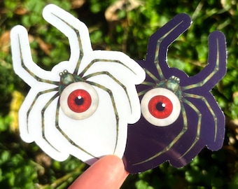 May include: Two spider stickers with red and white eyeballs. One spider is white with green legs, and the other is purple with green legs. The stickers are held up against a green background.