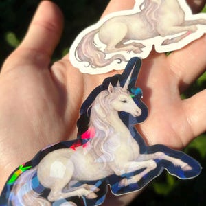 May include: Two unicorn stickers. One sticker features a white unicorn with a flowing mane and tail, lying down. The other sticker has a similar design, with a holographic, geometric border. The stickers are held in a hand.