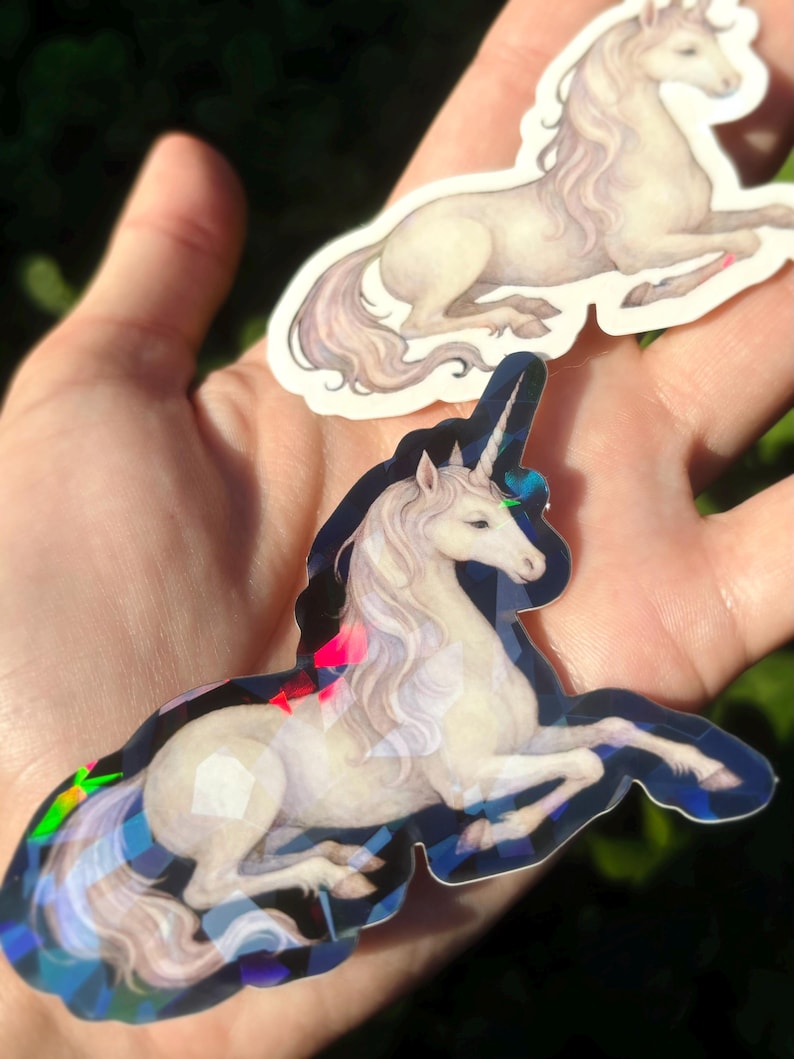 May include: Two unicorn stickers. One sticker features a white unicorn with a flowing mane and tail, lying down. The other sticker has a similar design, with a holographic, geometric border. The stickers are held in a hand.