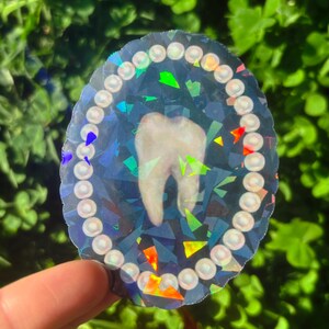 May include: A holographic sticker with a tooth design. The tooth is in the centre of an oval shape, encircled by a border of small, white, pearl-like beads. The background has a geometric, fractured pattern with iridescent colour accents.