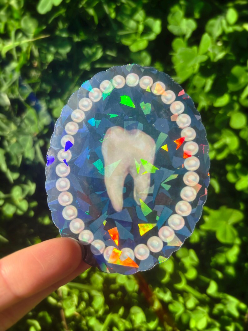 May include: A holographic sticker with a tooth design. The tooth is in the centre of an oval shape, encircled by a border of small, white, pearl-like beads. The background has a geometric, fractured pattern with iridescent colour accents.