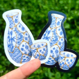 May include: Two die-cut stickers featuring a blue and white sake set with gold kintsugi-style cracks. The design includes floral, bamboo, and wave patterns on the bottle and cup. One sticker has a white border, while the other has a black border.